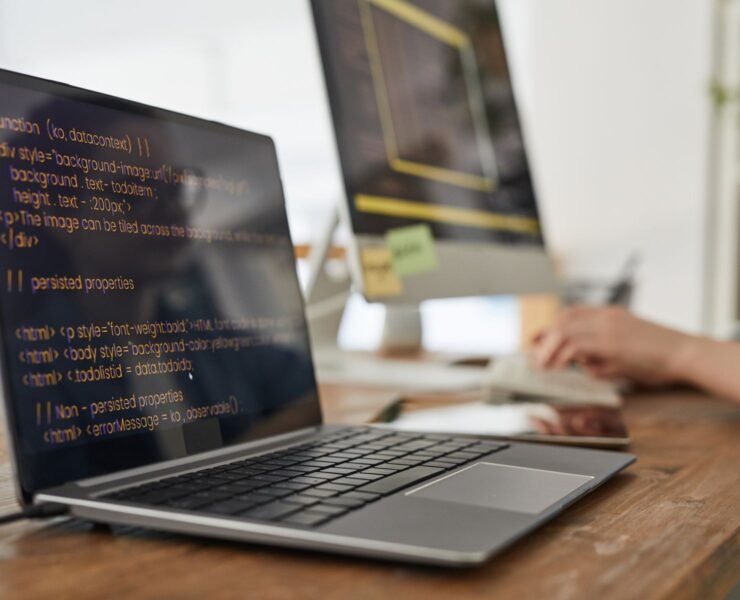 Close up of two computers with programming code on screen in minimal home office interior with hands typing in background, copy space