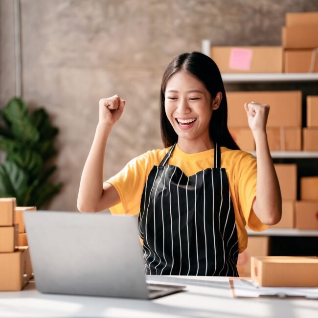 Small business parcel delivery concept, Female entrepreneur checking online order on laptop and raising arms to celebrate for work successfully while working about shopping online sales business.